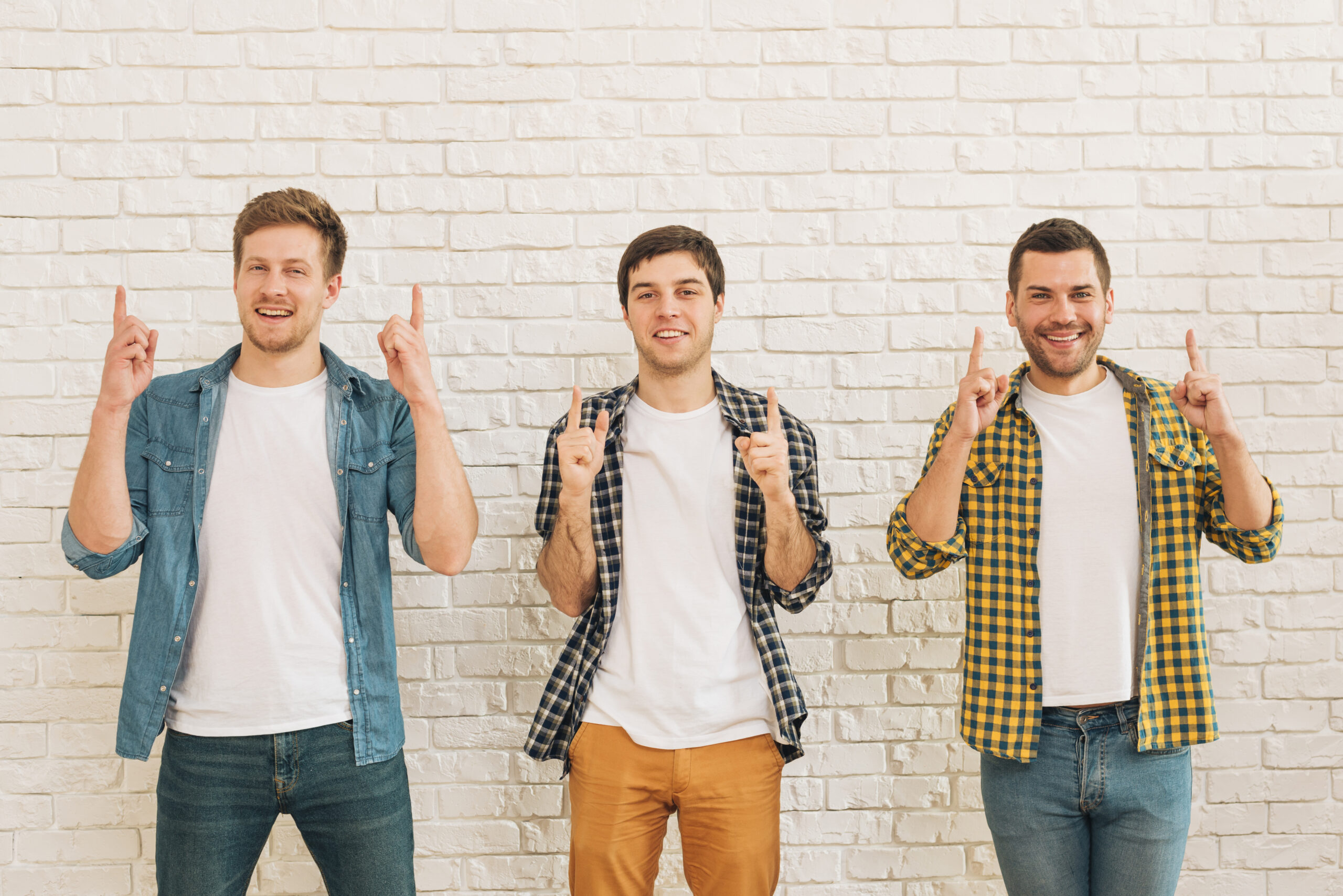 three-male-friends-standing-against-white-wall-pointing-finger-upward-looking-camera