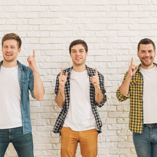 three-male-friends-standing-against-white-wall-pointing-finger-upward-looking-camera